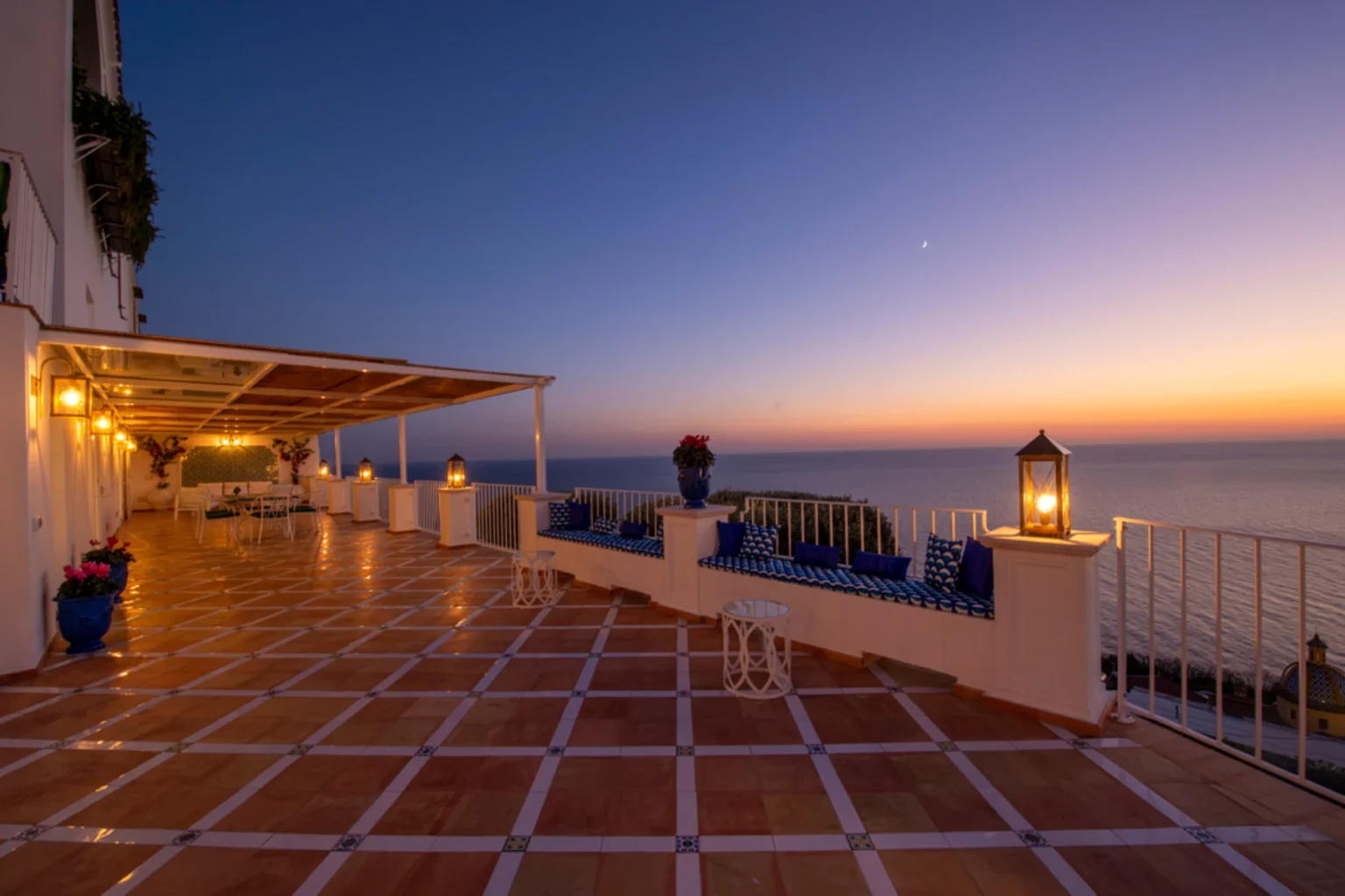 Scenic terrace with ocean view at sunset, featuring tiled flooring, decorative lanterns, and comfortable seating. The warm glow of lights enhances the tranquil atmosphere, perfect for relaxation or dining outdoors. | Only on SopranoVillas