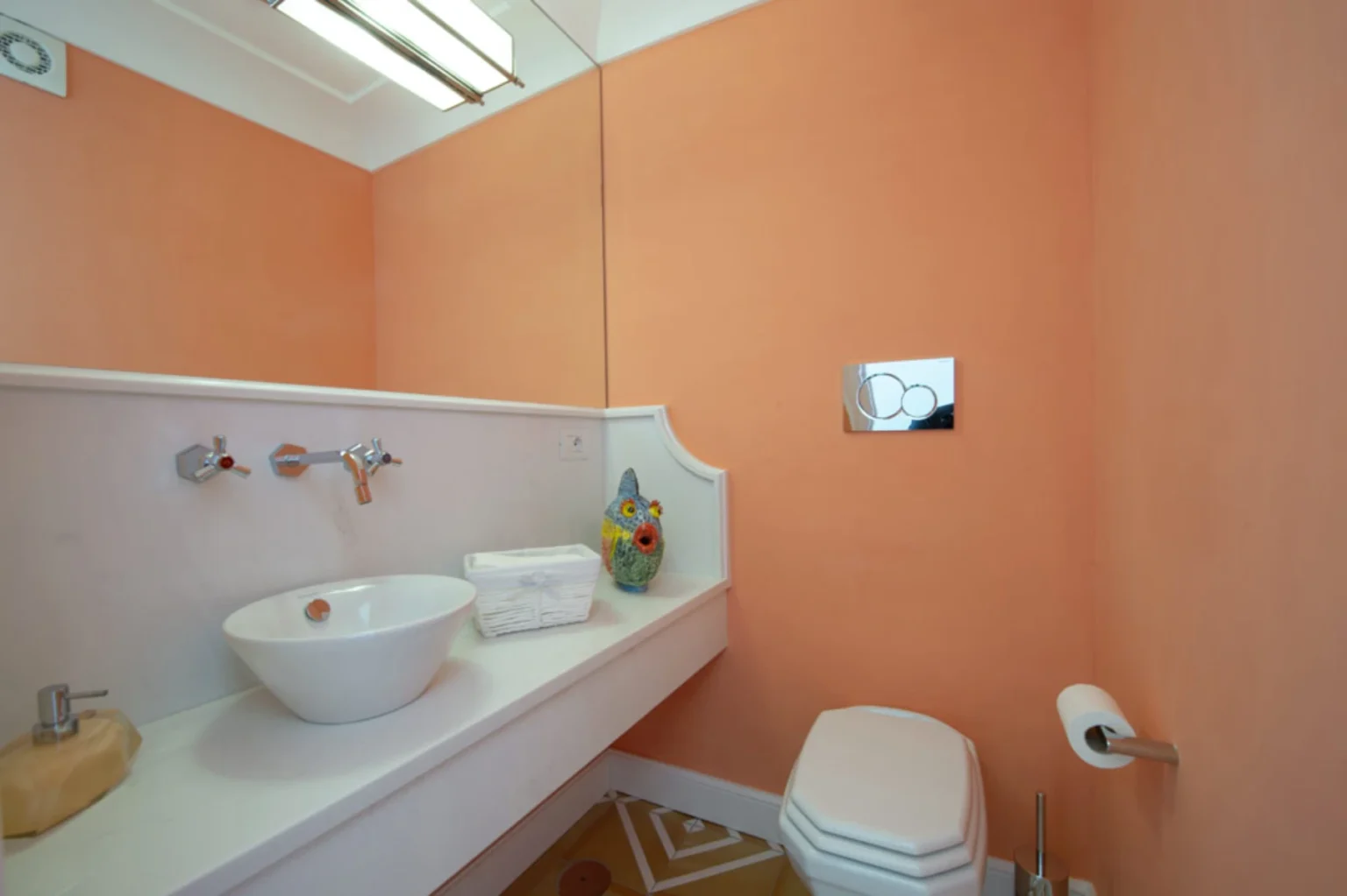 Modern bathroom with peach walls, a stylish white sink, and a decorative fish ornament. Features include a sleek toilet, a towel holder, and contemporary lighting, creating a bright and inviting atmosphere. | Only on SopranoVillas