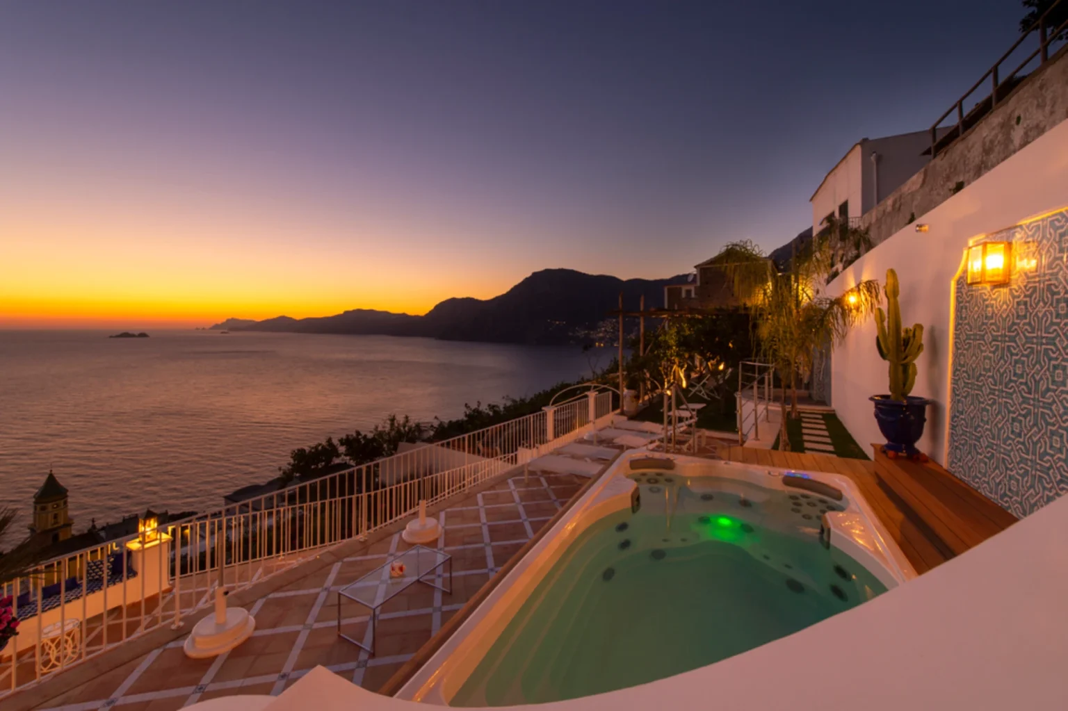 Scenic view of a luxury villa terrace overlooking the ocean at sunset, featuring a hot tub, mountain backdrop, and vibrant sky. Ideal for vacation rentals or travel destinations. | Only on SopranoVillas