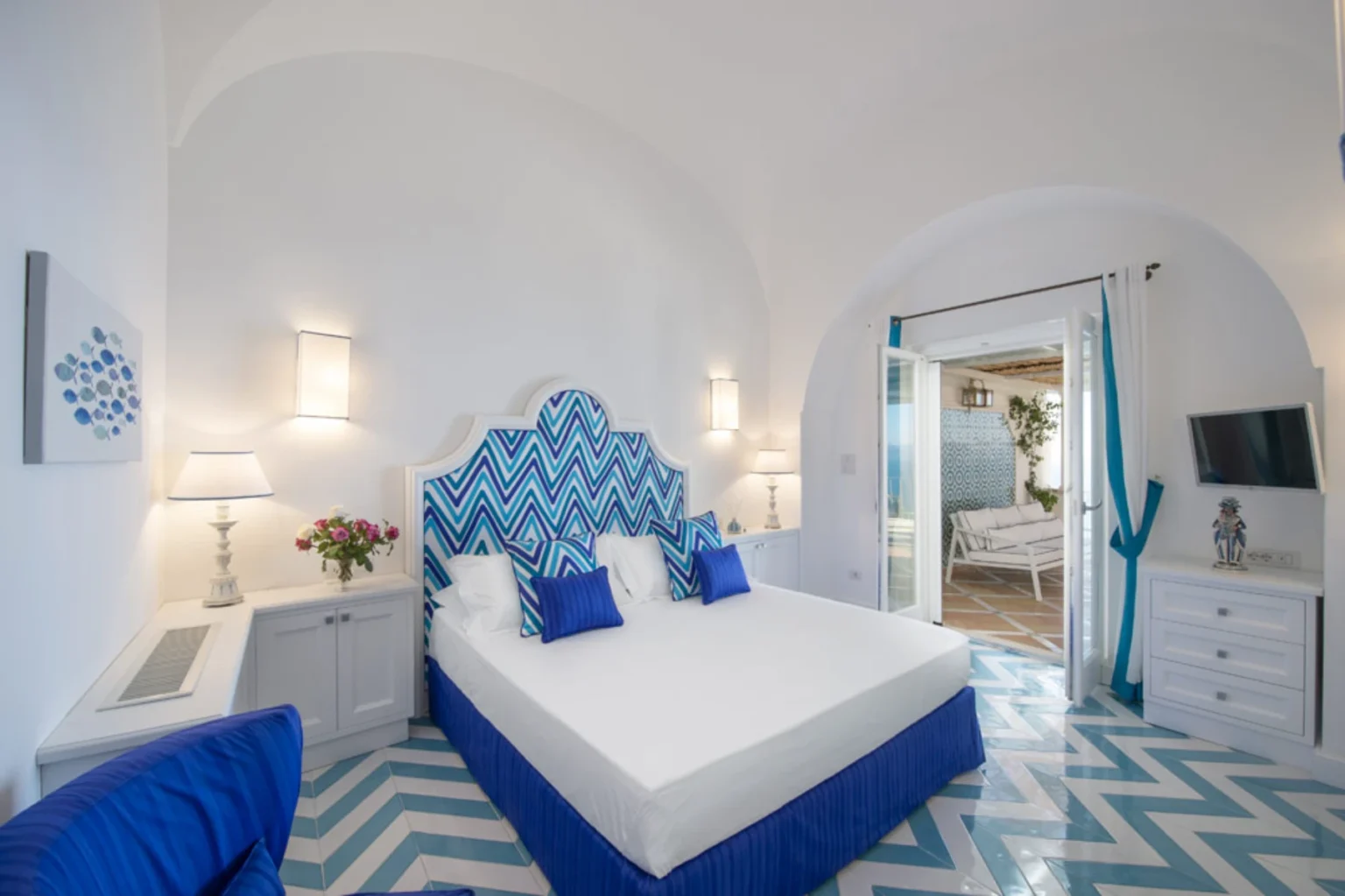 Bright and modern hotel room featuring a king-sized bed with a blue and white chevron headboard, accent pillows, and stylish decor. The room includes a cozy seating area, a flat-screen TV, and an entrance to a scenic outdoor space. Elegant lighting and a floral arrangement enhance the inviting atmosphere. | Only on SopranoVillas