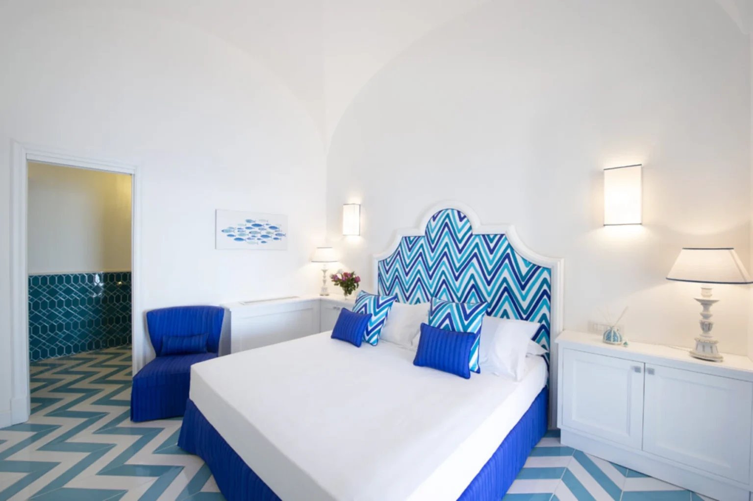 Bright and stylish bedroom featuring a white bed with blue and turquoise chevron-patterned headboard, complemented by matching decorative pillows. The room includes a blue accent chair, a white nightstand with a lamp, and a vibrant tiled floor. The walls are painted white, enhancing the airy and modern aesthetic. Perfect for a coastal-themed interior design. | Only on SopranoVillas