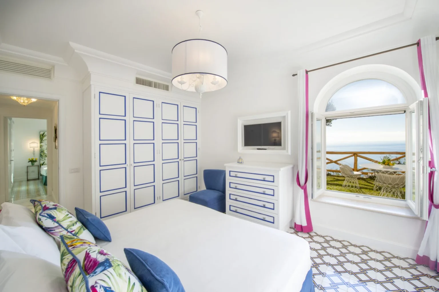 Bright and airy bedroom featuring a white bed with colorful pillows, a stylish blue armchair, and a modern dresser. The room has a large window with scenic views of the ocean and outdoor seating. Decorated with blue accents and elegant light fixtures, this space combines comfort and aesthetic appeal. | Only on SopranoVillas