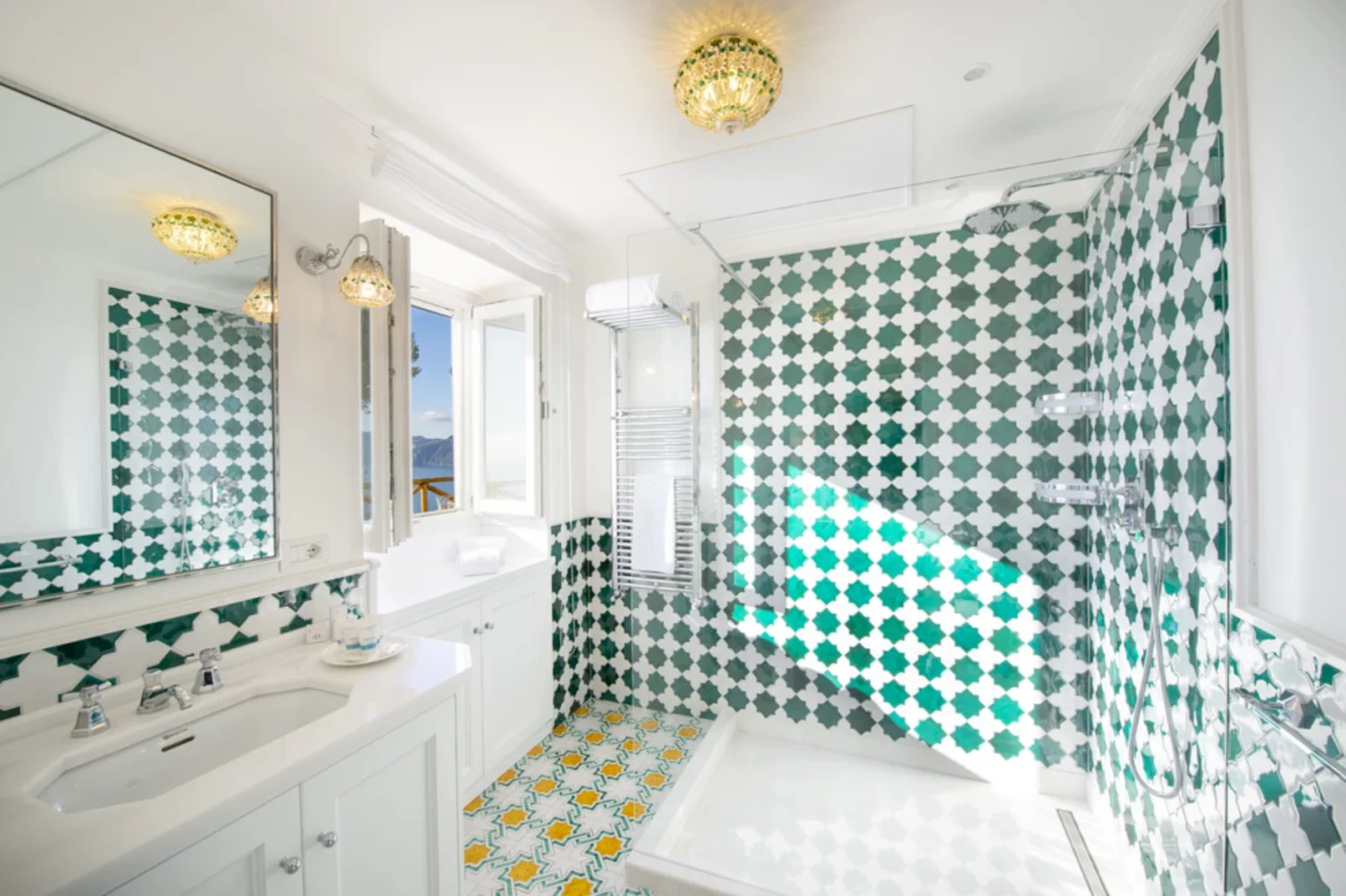 Modern bathroom featuring green and white patterned tiles, a spacious shower area, a double sink vanity, and a large mirror. Natural light floods the space through open windows, creating a bright and airy atmosphere. | Only on SopranoVillas