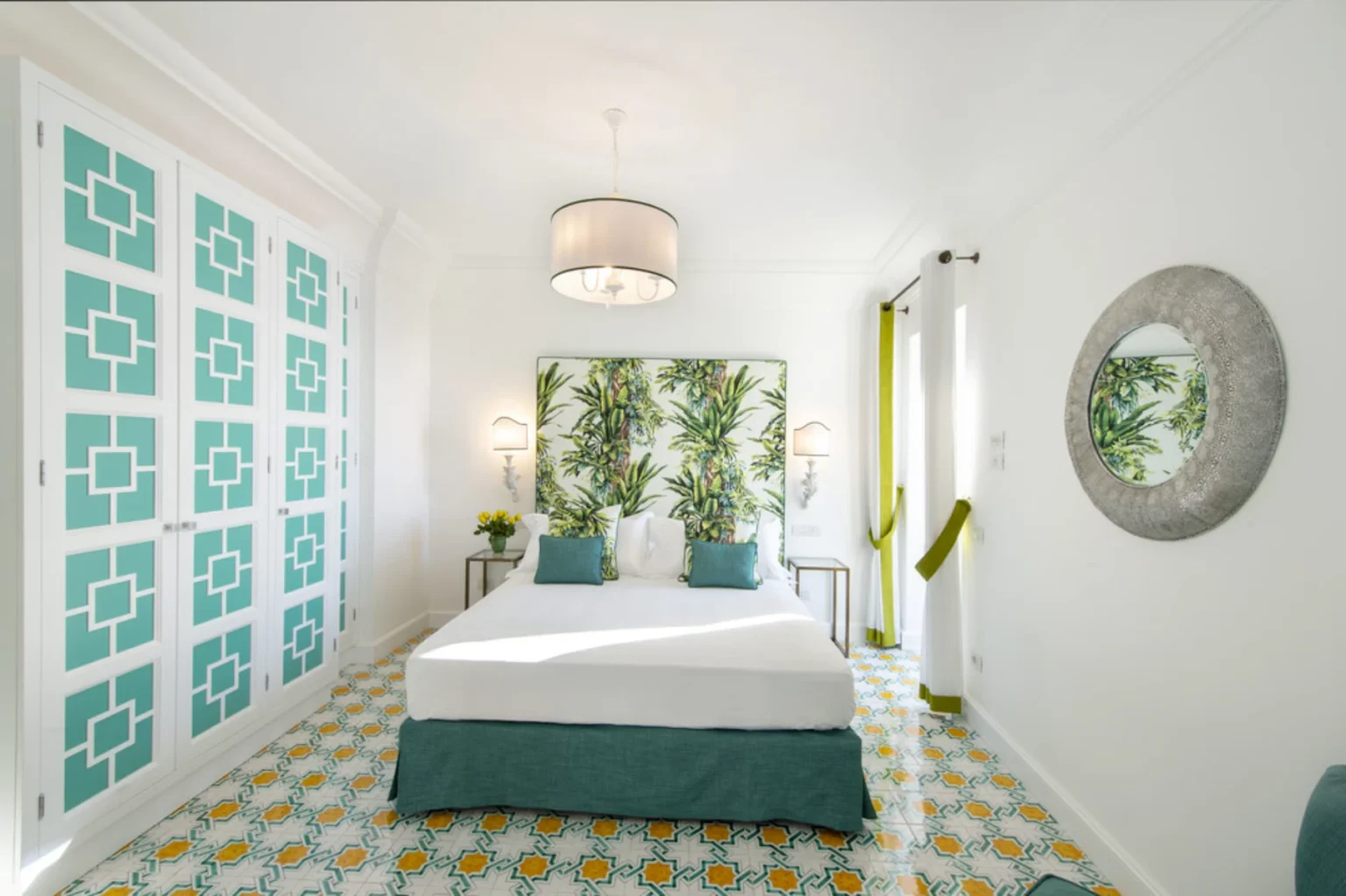 Bright and airy bedroom featuring a large bed with teal accents, a tropical-themed headboard, and stylish patterned flooring. The room includes white cabinetry with teal designs, contemporary lighting, and decorative elements like a round mirror and fresh flowers, creating a modern yet relaxed atmosphere. | Only on SopranoVillas