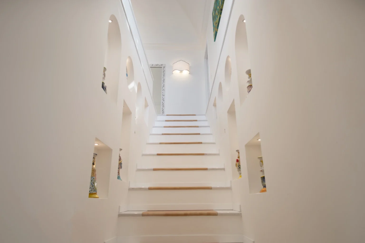 Modern staircase with white walls and wooden accents, featuring decorative alcoves showcasing colorful figurines. Bright, airy design with natural light illuminating the space. Ideal for home decor inspiration. | Only on SopranoVillas