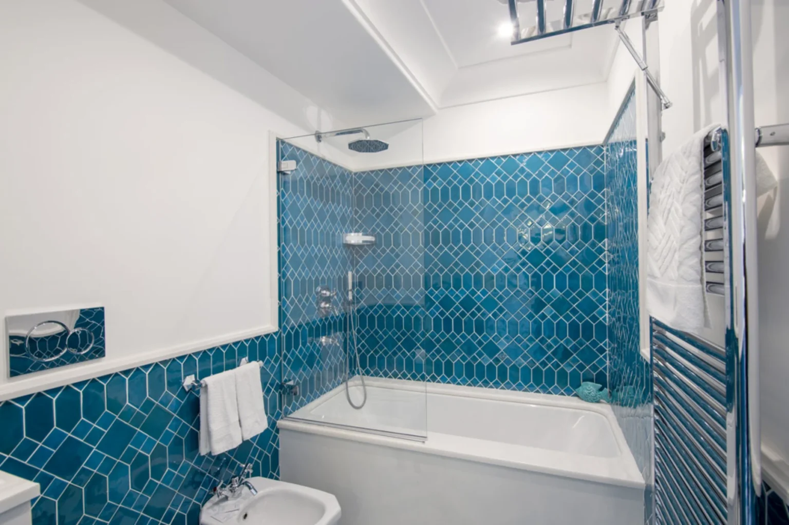 Modern bathroom featuring a stylish blue hexagonal tile design, a glass shower enclosure, a white bathtub, and chrome towel radiator with neatly hung towels. | Only on SopranoVillas