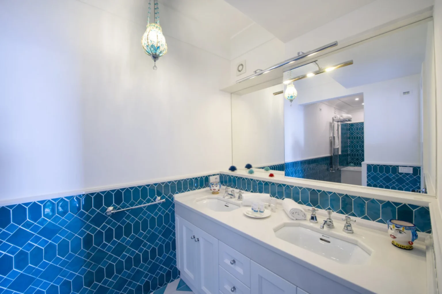 Modern bathroom featuring a double vanity with white cabinetry, a blue hexagonal tile backsplash, and a stylish pendant light. The space is illuminated by natural light and includes essential toiletries, fresh towels, and a mirror reflecting the contemporary design. | Only on SopranoVillas