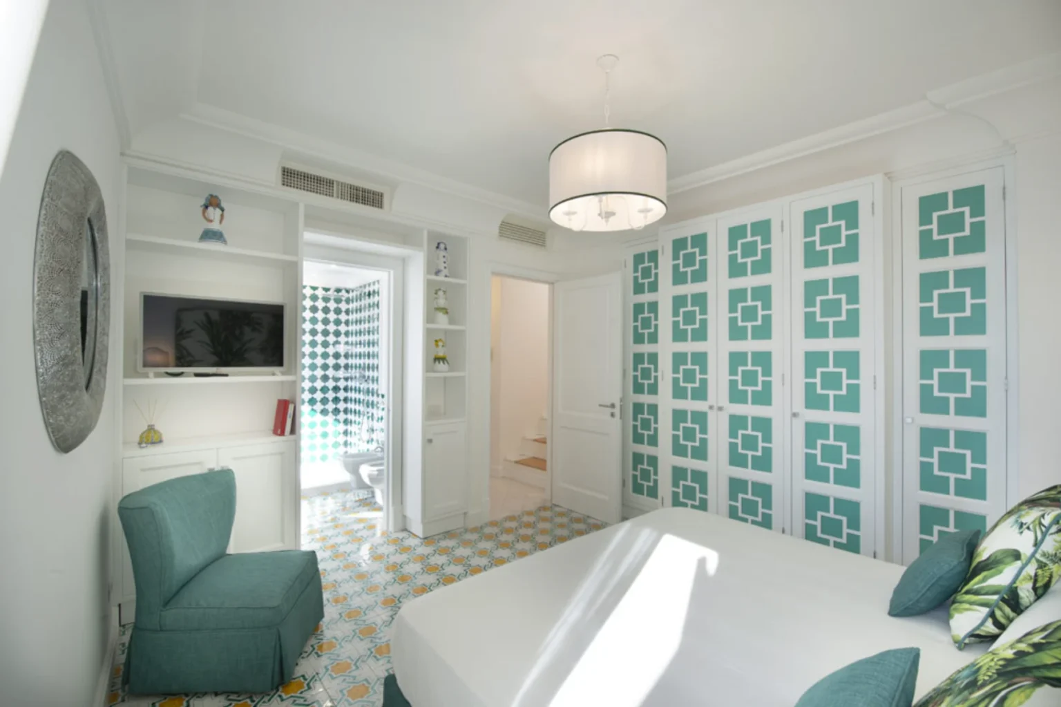 Bright and modern bedroom featuring a white bed, teal accent chair, and geometric patterned wardrobe. The room includes a wall-mounted TV and vibrant tiled flooring, with a glimpse of a bathroom in the background. Ideal for a stylish and comfortable stay. | Only on SopranoVillas