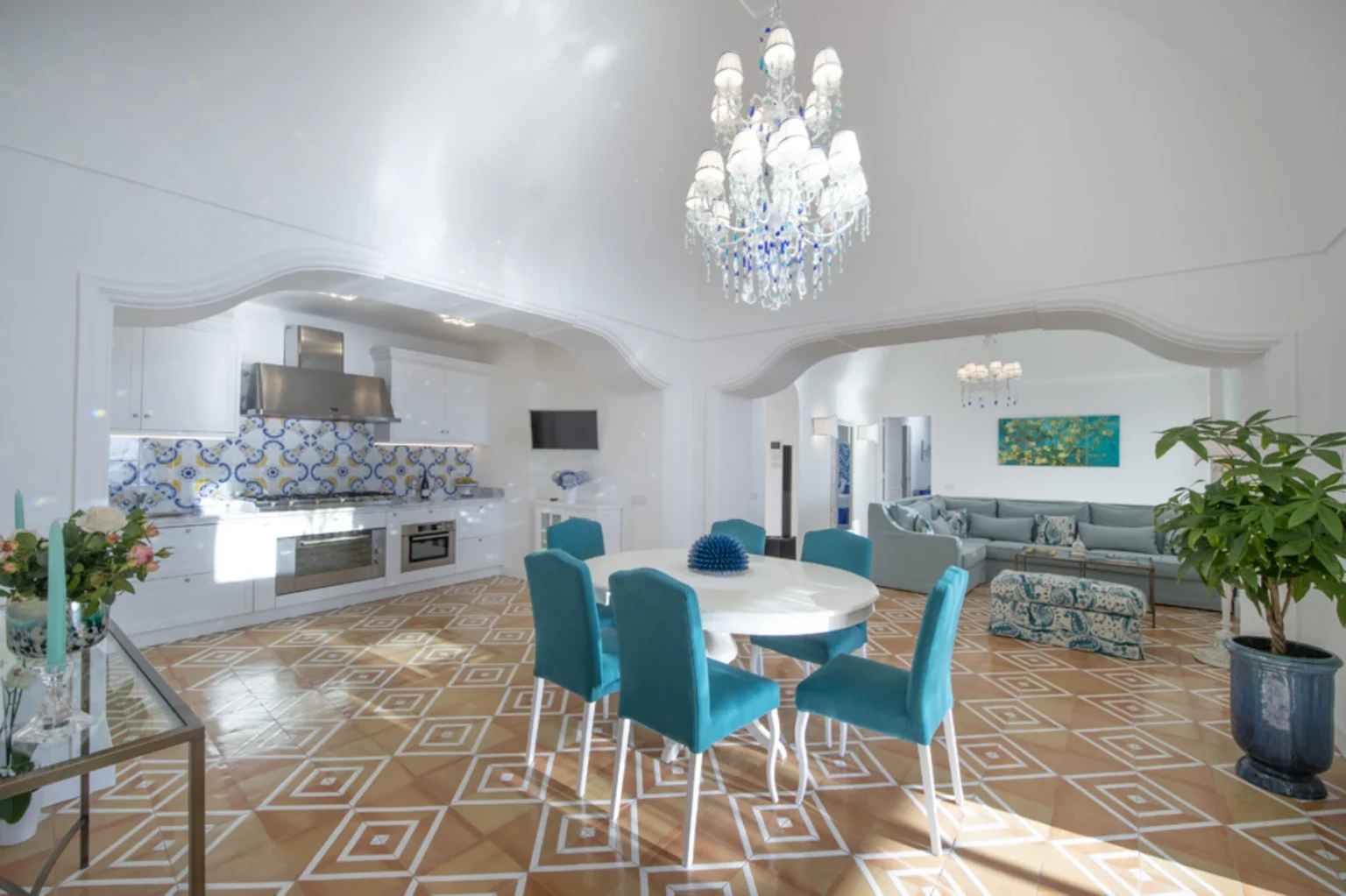 Bright and modern dining area featuring a round white table surrounded by turquoise chairs, with a stylish chandelier overhead. The kitchen showcases a blue and white tiled backsplash, stainless steel appliances, and elegant decor. The room boasts a spacious layout with geometric patterned flooring and a cozy living area visible in the background. | Only on SopranoVillas