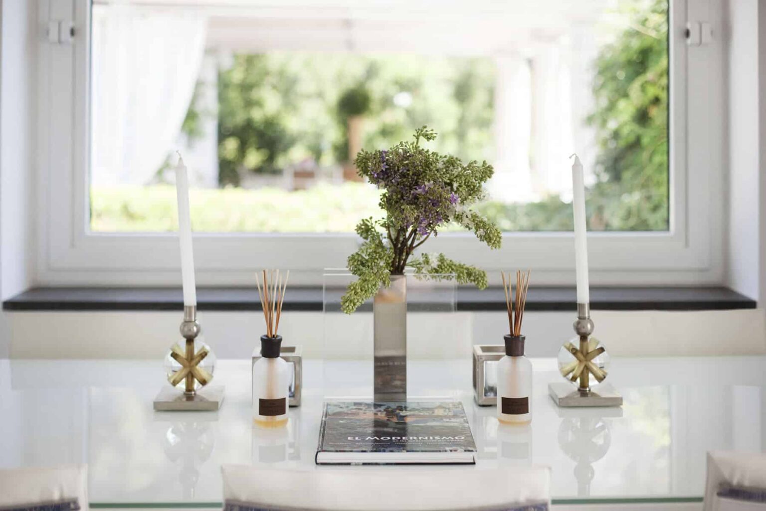 Modern dining table centerpiece featuring a vase with greenery, two decorative candle holders, and scented oil bottles, set against a bright window view. | Only on SopranoVillas