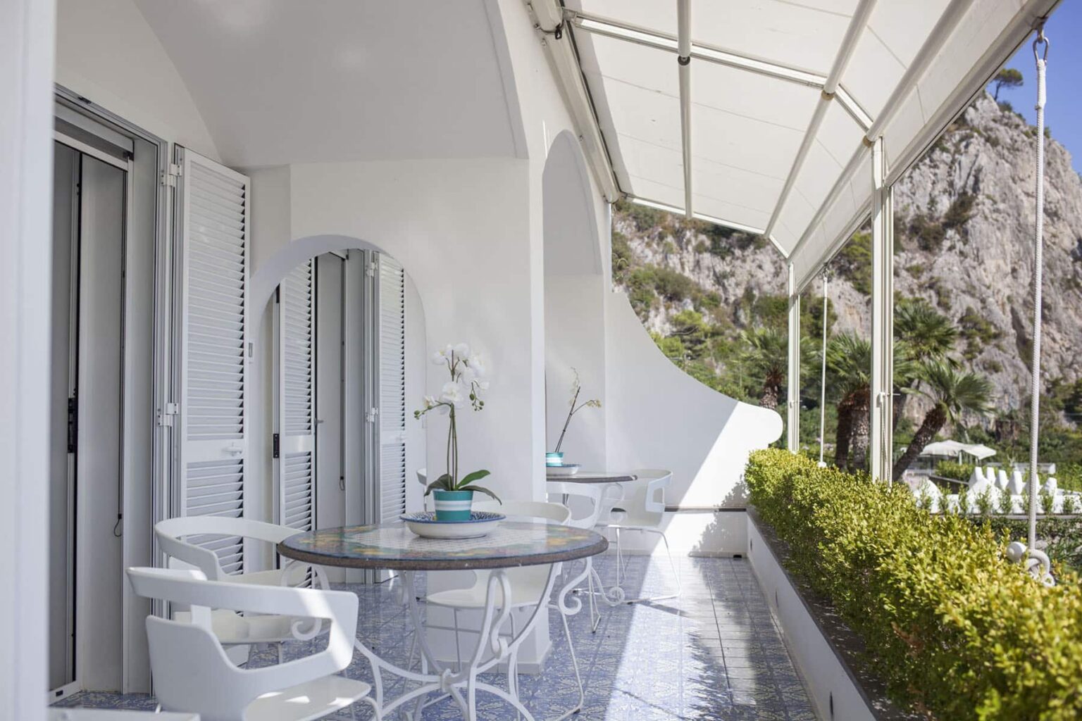 Modern balcony with white furniture, a round table, and a potted plant, featuring a scenic view of greenery and rocky landscape under a shaded awning. Ideal for relaxation and outdoor dining. | Only on SopranoVillas