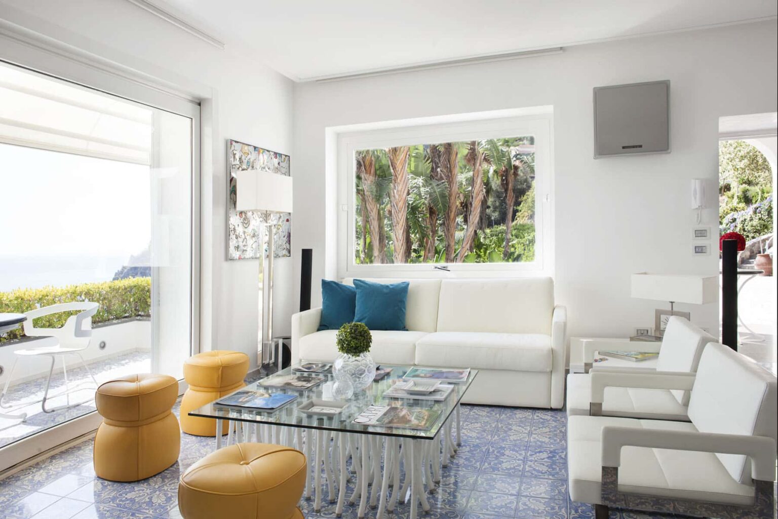 Modern living room with white furniture, colorful accent stools, and a glass coffee table, featuring large windows that offer a view of greenery outside. Bright and airy atmosphere with contemporary decor elements. | Only on SopranoVillas