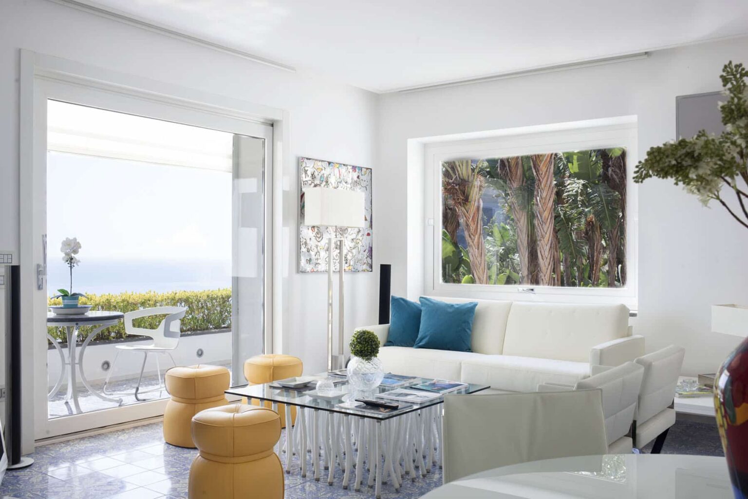 Bright and modern living room featuring a white sofa, glass coffee table, and yellow accent stools, with large windows showcasing a scenic ocean view and greenery outside. | Only on SopranoVillas