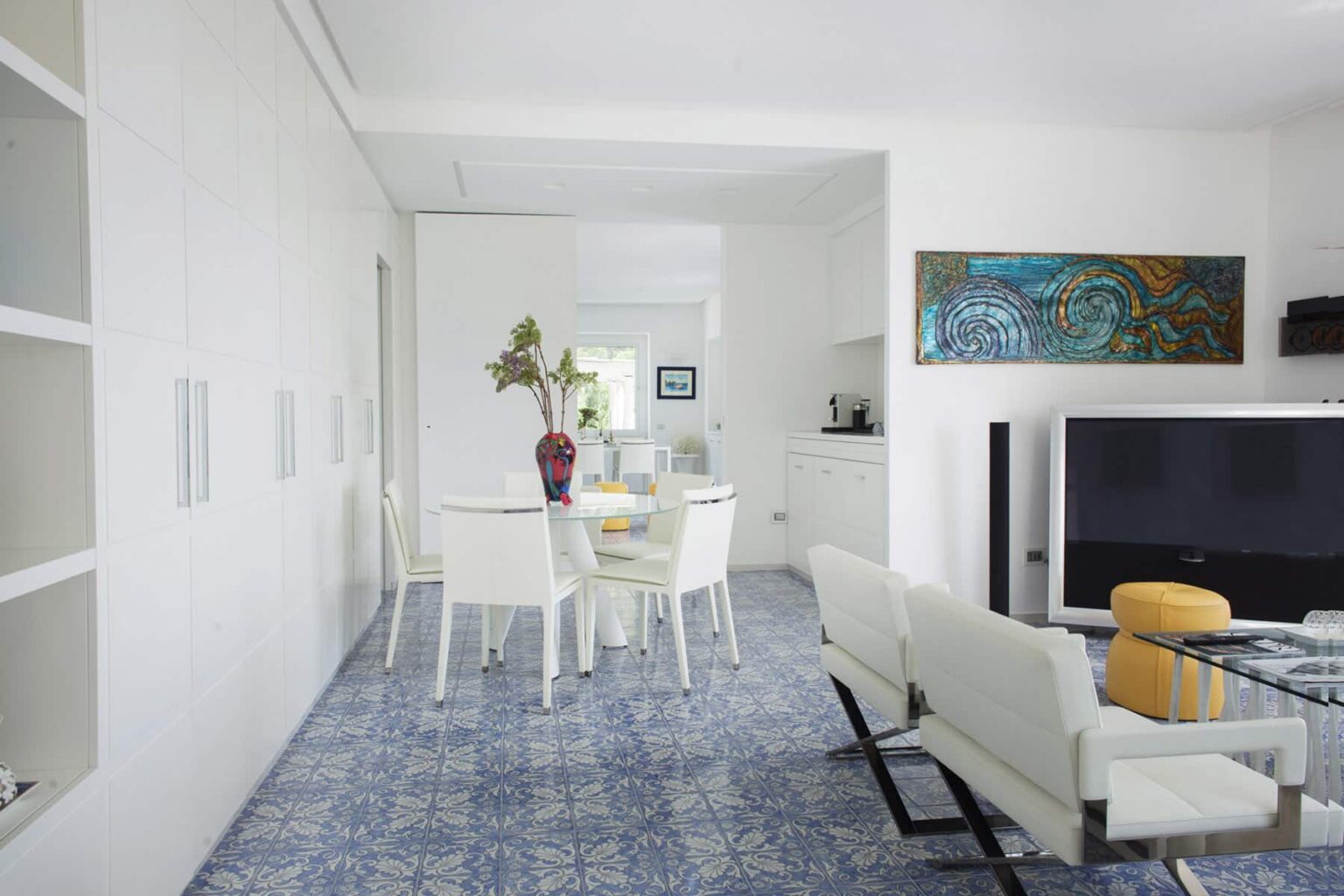 Bright and airy modern living space featuring a white dining table surrounded by stylish white chairs, a colorful vase with flowers, and decorative art on the walls. The blue patterned tile floor adds a touch of elegance, complemented by a sleek television and contemporary furniture. | Only on SopranoVillas
