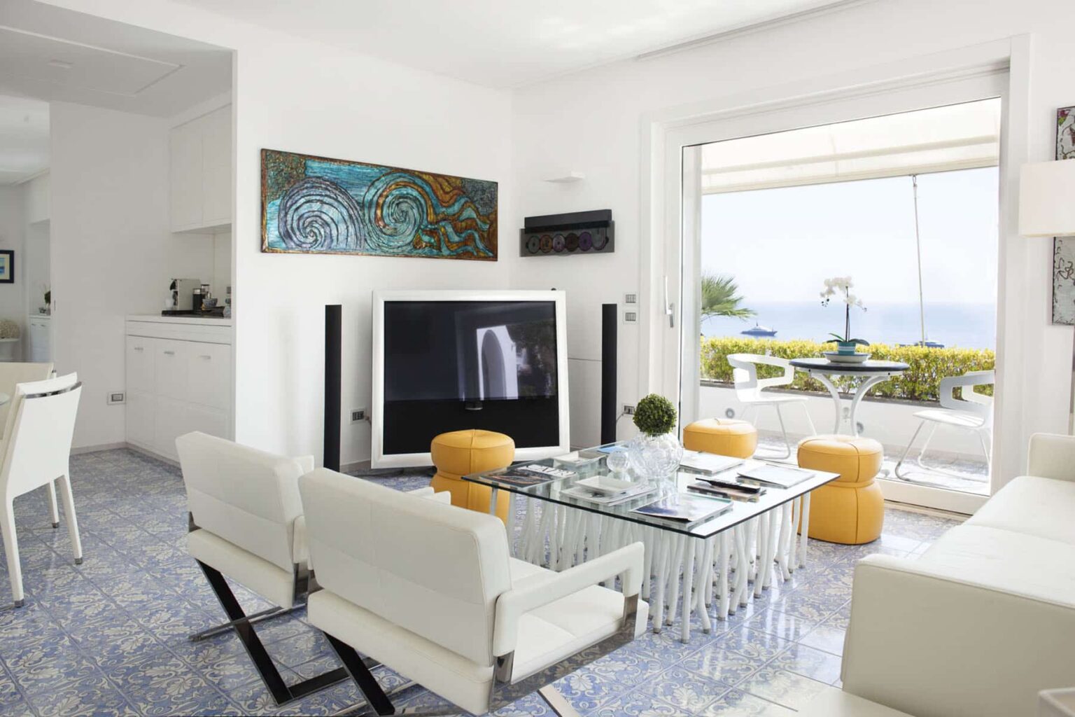 Modern living room with white furniture, a glass coffee table, and a large flat-screen TV, featuring a scenic view through glass doors leading to a balcony. Bright and airy atmosphere with decorative art on the walls. | Only on SopranoVillas