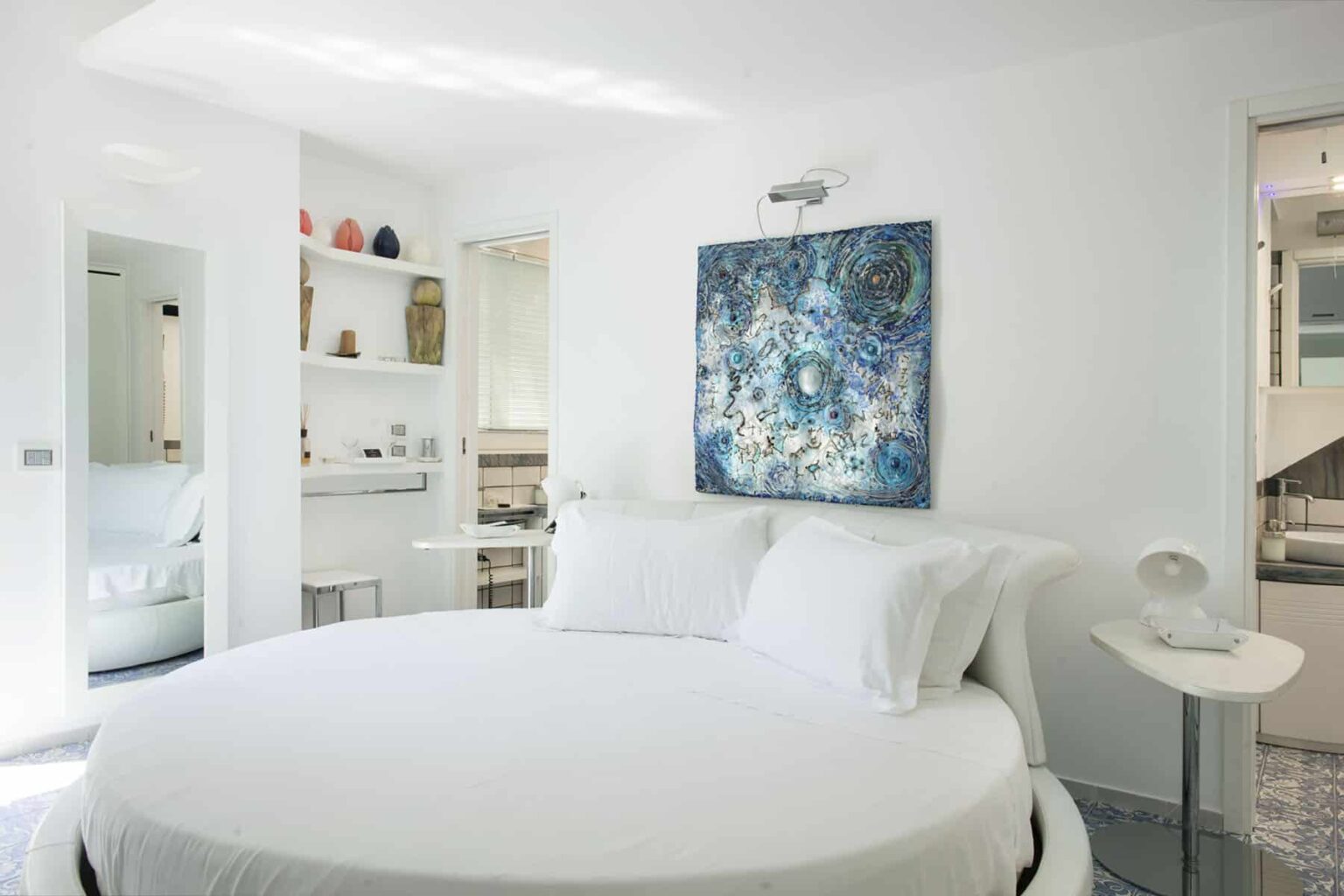 Modern bedroom featuring a round white bed with plush pillows, a vibrant abstract painting in blue tones on the wall, and minimalist decor. The room includes a small side table, a sleek lamp, and a mirror reflecting additional furnishings, creating a bright and airy atmosphere. | Only on SopranoVillas