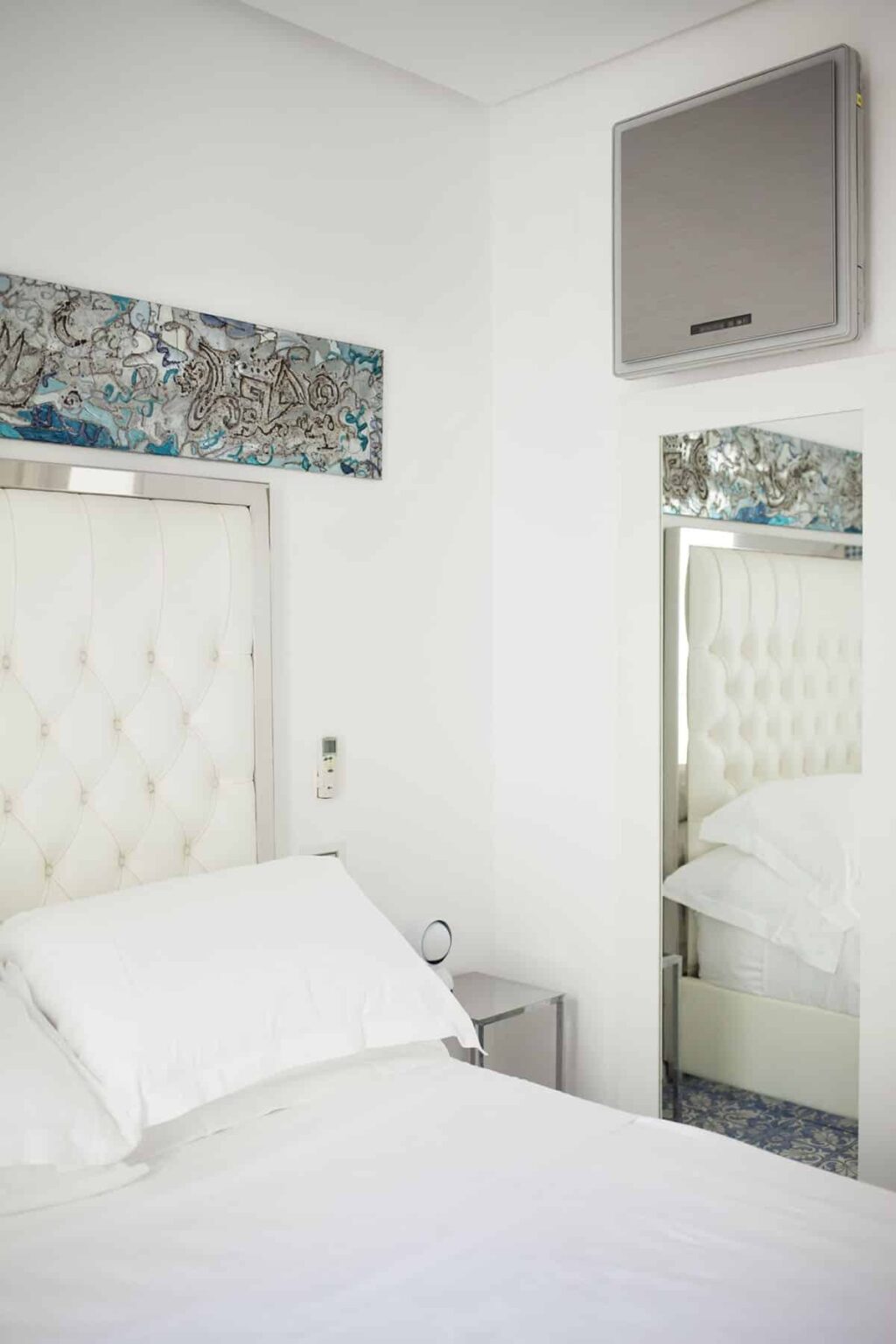 Elegant bedroom interior featuring a tufted white headboard, soft bedding, and a decorative mirror. The walls are adorned with a colorful artwork, creating a bright and inviting atmosphere. | Only on SopranoVillas