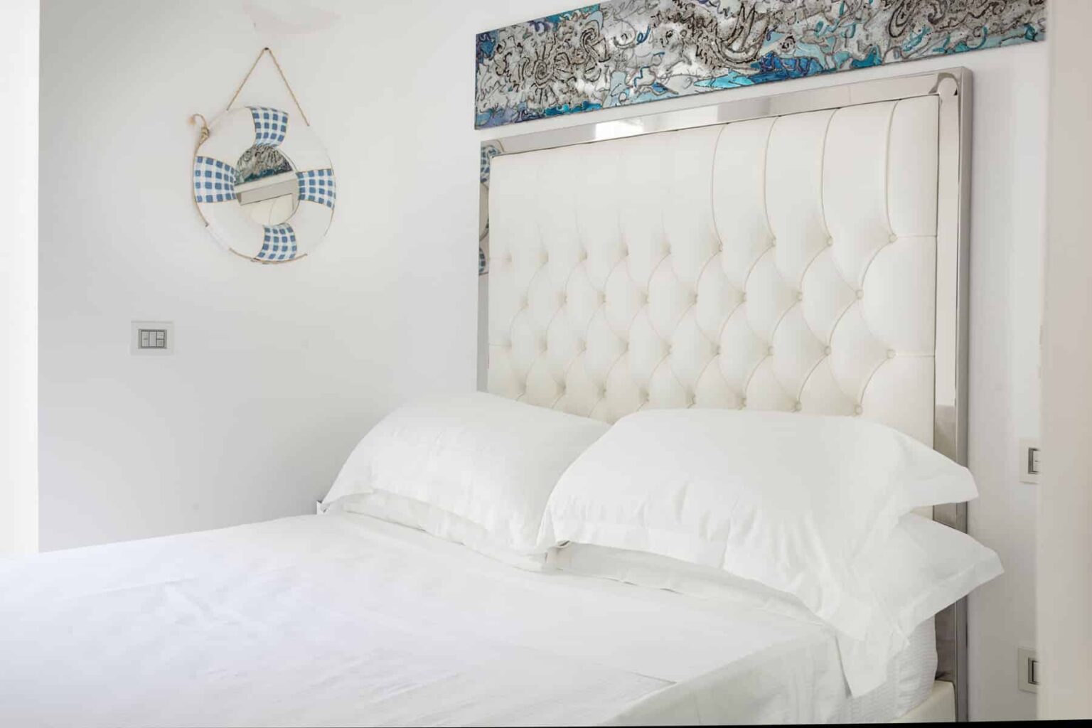 Modern bedroom interior featuring a plush, tufted white headboard with two fluffy white pillows on a neatly made bed. A decorative life preserver with blue accents hangs on the wall, complemented by a colorful artwork above the headboard. The room exudes a fresh and airy atmosphere with a minimalist design. | Only on SopranoVillas