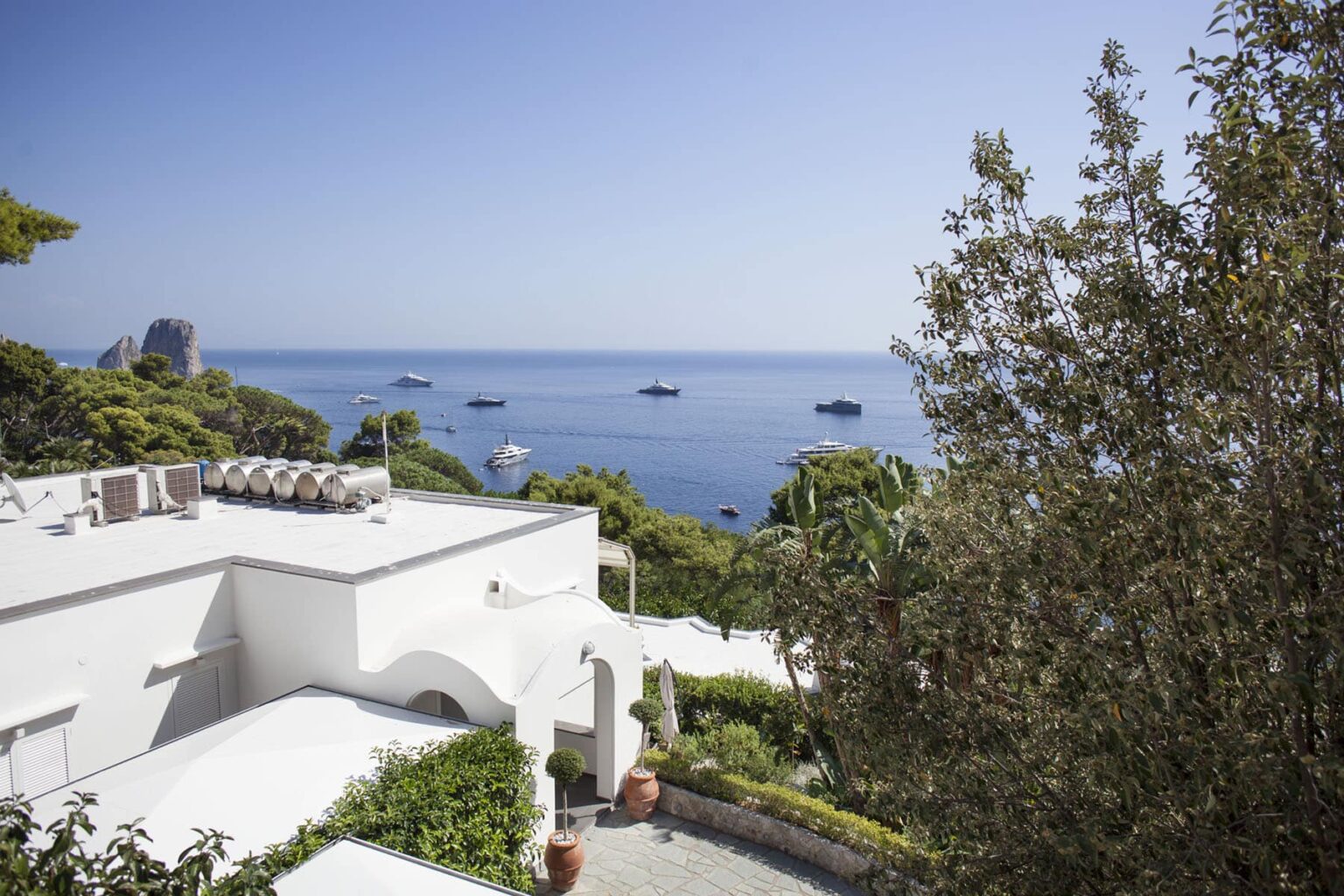 Scenic view of a coastal villa overlooking a tranquil sea with several boats in the distance, surrounded by lush greenery and clear blue skies. | Only on SopranoVillas