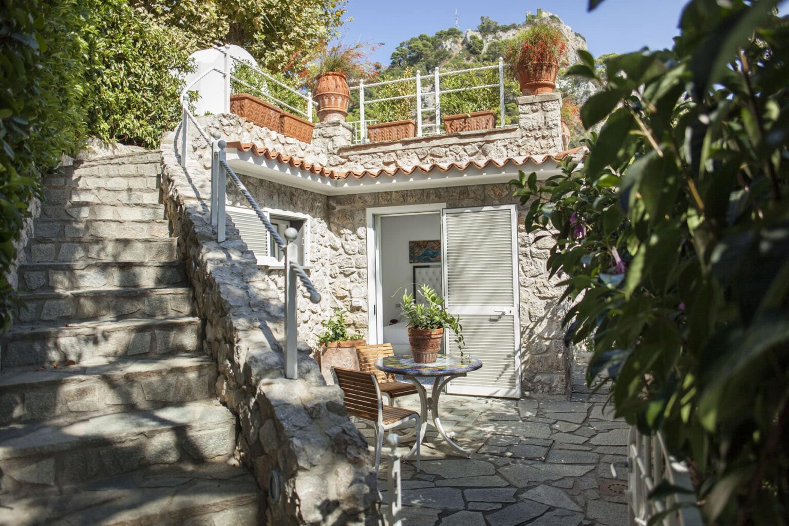 Charming stone cottage with a terrace and potted plants, surrounded by greenery and stairs leading to the entrance, showcasing a serene outdoor setting. | Only on SopranoVillas