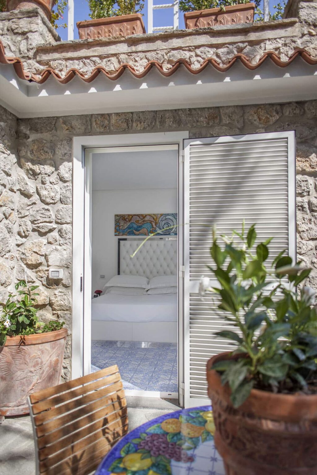 Cozy bedroom with a white bed and decorative artwork, viewed through a modern sliding glass door, framed by a natural stone wall and lush green plants. | Only on SopranoVillas