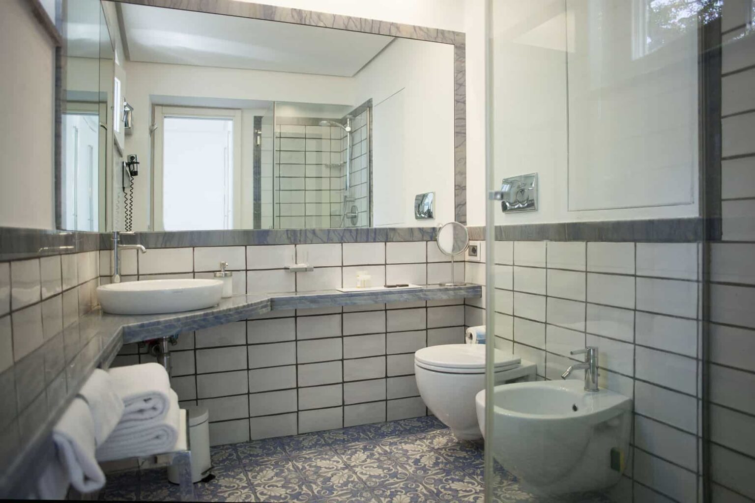 Modern bathroom with gray and white tile design, featuring a large mirror, sink, bidet, and toilet, complemented by natural light from a window. | Only on SopranoVillas