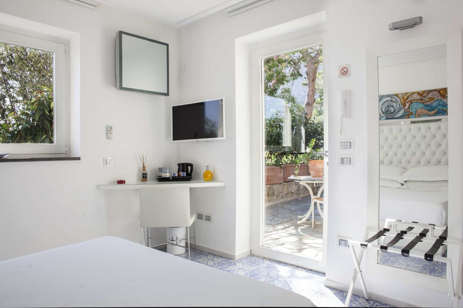 Bright and modern hotel room featuring a comfortable bed, a small workspace with a TV, and large windows that open to a private outdoor patio with seating. The minimalist design includes white walls and contemporary furnishings, creating a serene atmosphere. | Only on SopranoVillas
