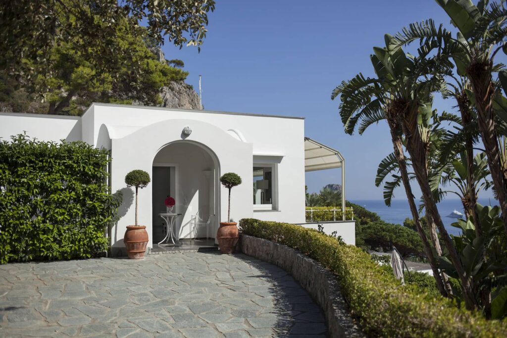 Modern white villa entrance surrounded by lush greenery and palm trees, featuring a stone pathway and scenic ocean view in the background. Ideal for luxury travel and vacation rentals. | Only on SopranoVillas