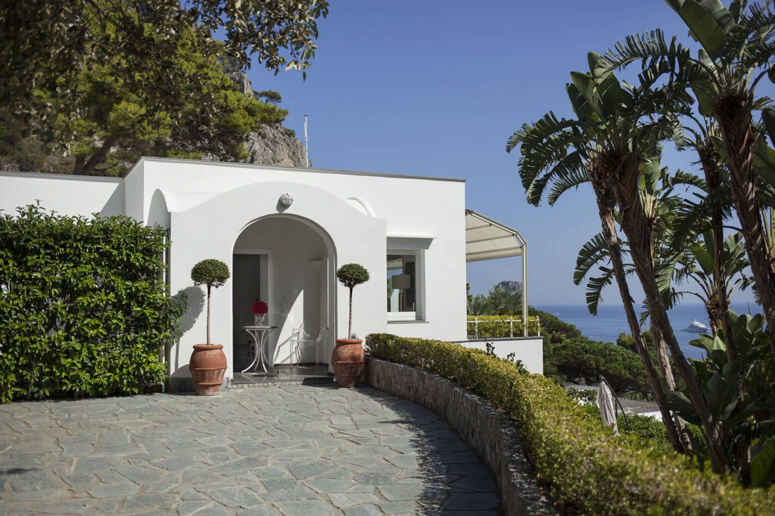 Modern white villa entrance surrounded by lush greenery and palm trees, featuring a stone pathway and scenic ocean view in the background. Ideal for luxury travel and vacation rentals. | Only on SopranoVillas