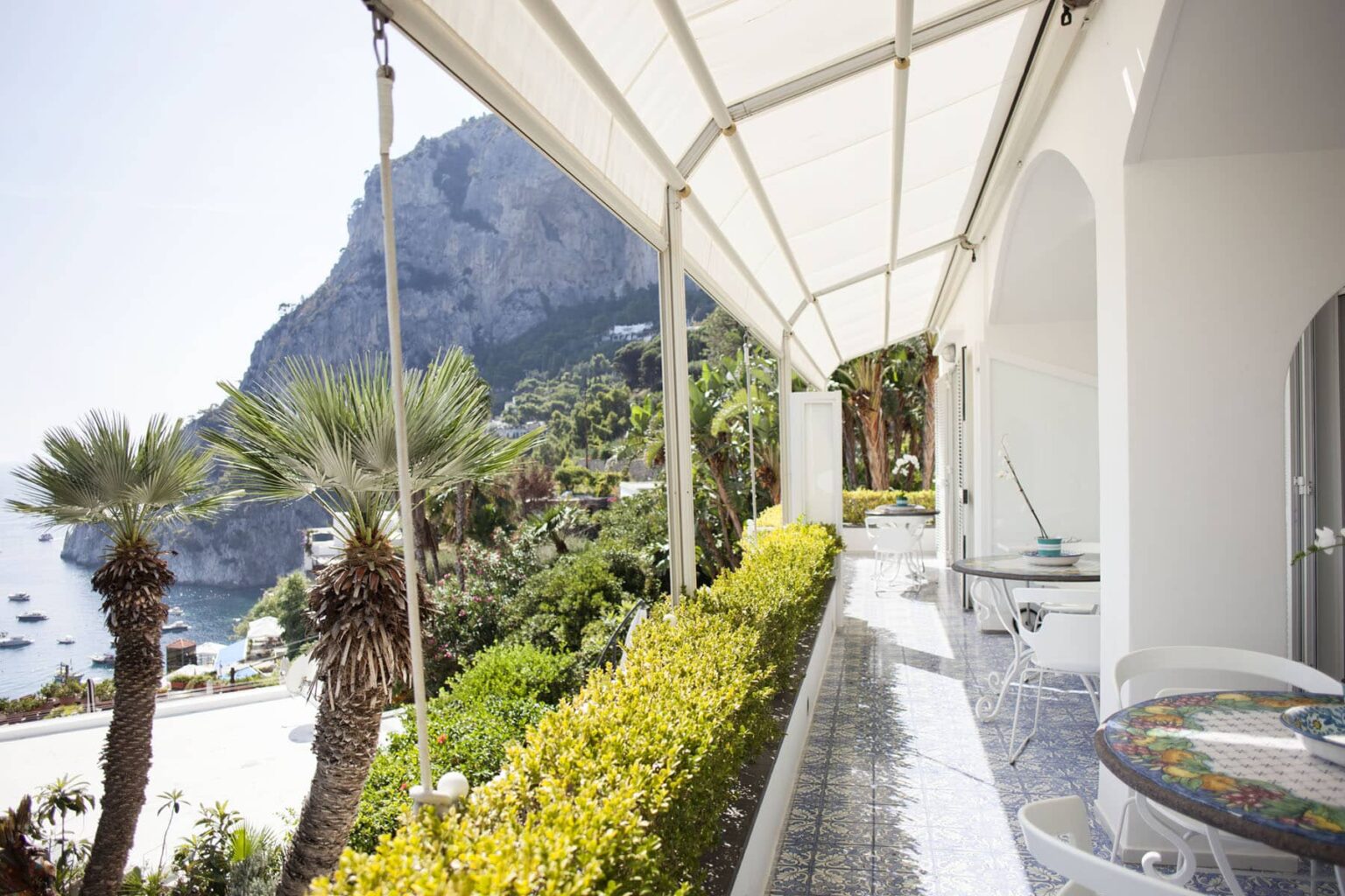 Scenic view of a coastal villa in Capri, featuring a terrace with modern furniture, lush greenery, and mountains in the background overlooking the sea. | Only on SopranoVillas