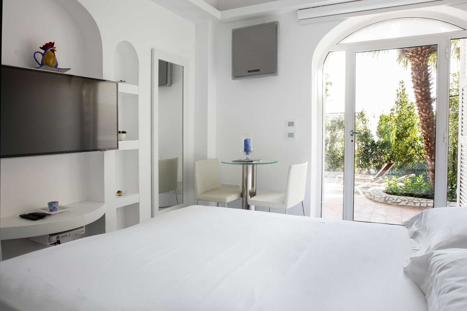 Modern hotel room featuring a white bed, flat-screen TV, and a glass dining table with chairs. Large windows open to a garden view, creating a bright and airy atmosphere. Ideal for relaxation and comfort during your stay. | Only on SopranoVillas