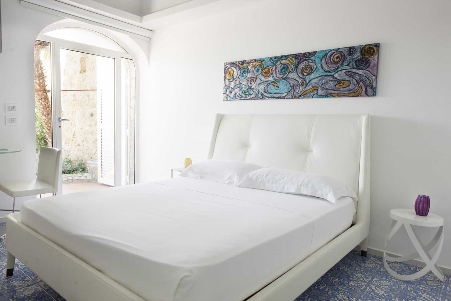 Modern bedroom featuring a white upholstered bed with crisp linens, a colorful abstract painting above, and natural light streaming in through an open door. The room has minimalistic decor with a small side table and a vibrant rug, creating a serene and inviting atmosphere. | Only on SopranoVillas