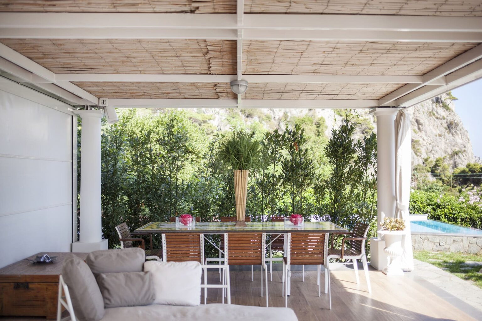 Outdoor dining area featuring a wooden table surrounded by white chairs, set under a shaded pergola. Lush greenery and a swimming pool can be seen in the background, creating a serene atmosphere for al fresco dining. | Only on SopranoVillas