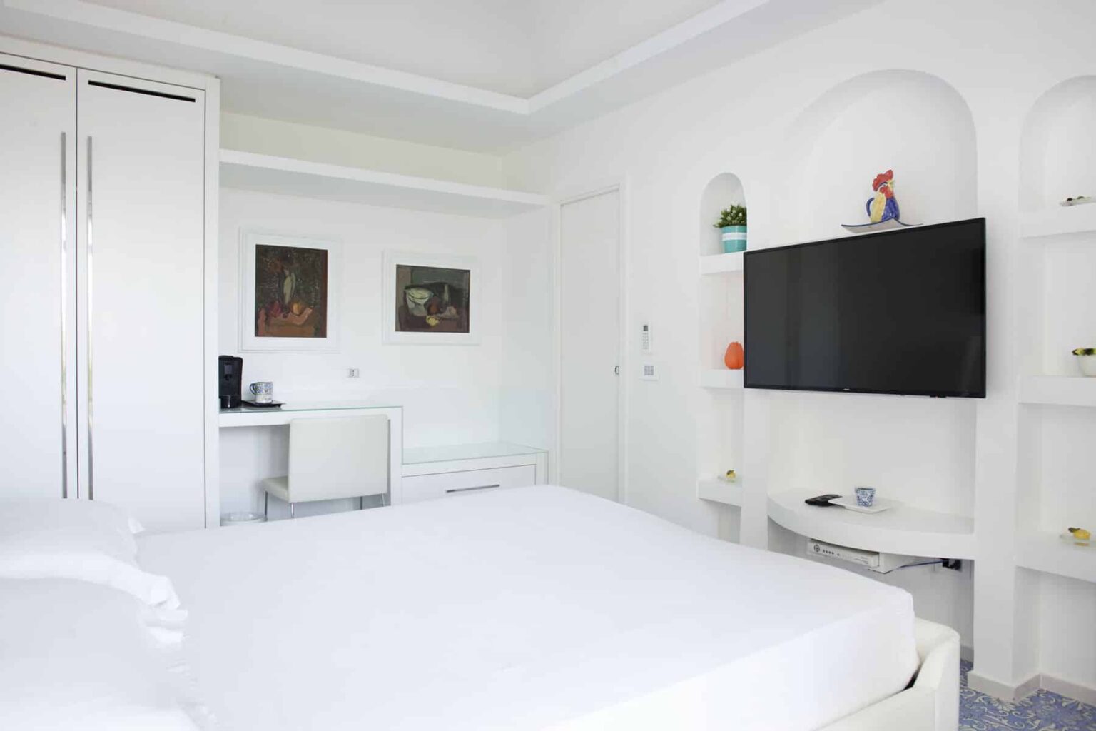 Modern white bedroom featuring a large bed, wall-mounted TV, and stylish storage solutions, complemented by decorative artwork and a minimalist design. | Only on SopranoVillas