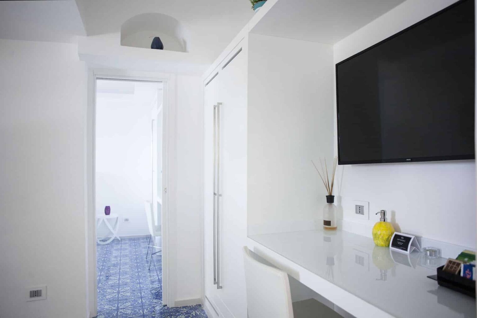 Modern hotel room interior featuring a sleek white design, a flat-screen TV mounted on the wall, and a minimalist workspace with decorative elements. The image captures a bright ambiance with a visible doorway leading to another area. | Only on SopranoVillas