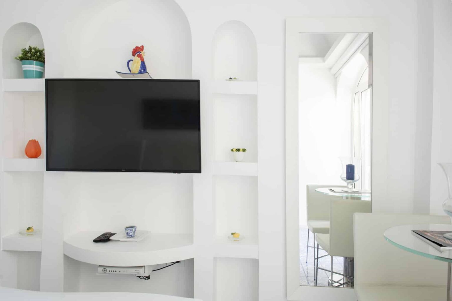 Modern living room with a mounted flat-screen TV, white shelving unit displaying decorative items, and a mirror reflecting a bright, airy space. | Only on SopranoVillas