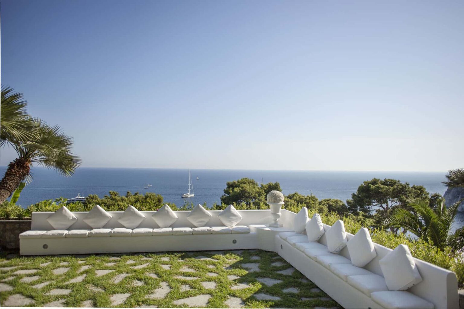 Luxurious outdoor seating area with white cushions overlooking a serene ocean view, surrounded by lush greenery and palm trees. Ideal for relaxation and enjoying coastal scenery. | Only on SopranoVillas