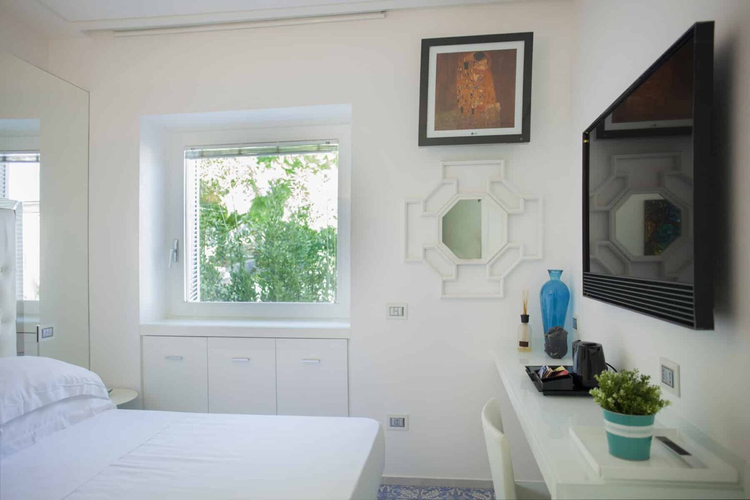 Bright and modern bedroom featuring a white bed, a large window with greenery outside, a stylish wall-mounted TV, and minimalistic decor including a small plant and artwork. | Only on SopranoVillas
