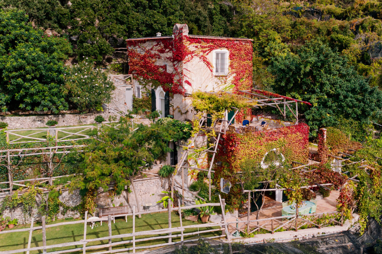Aerial view of a charming rustic house covered in vibrant red vines, surrounded by lush greenery and terraced gardens, showcasing a serene outdoor space ideal for relaxation. | Only on SopranoVillas