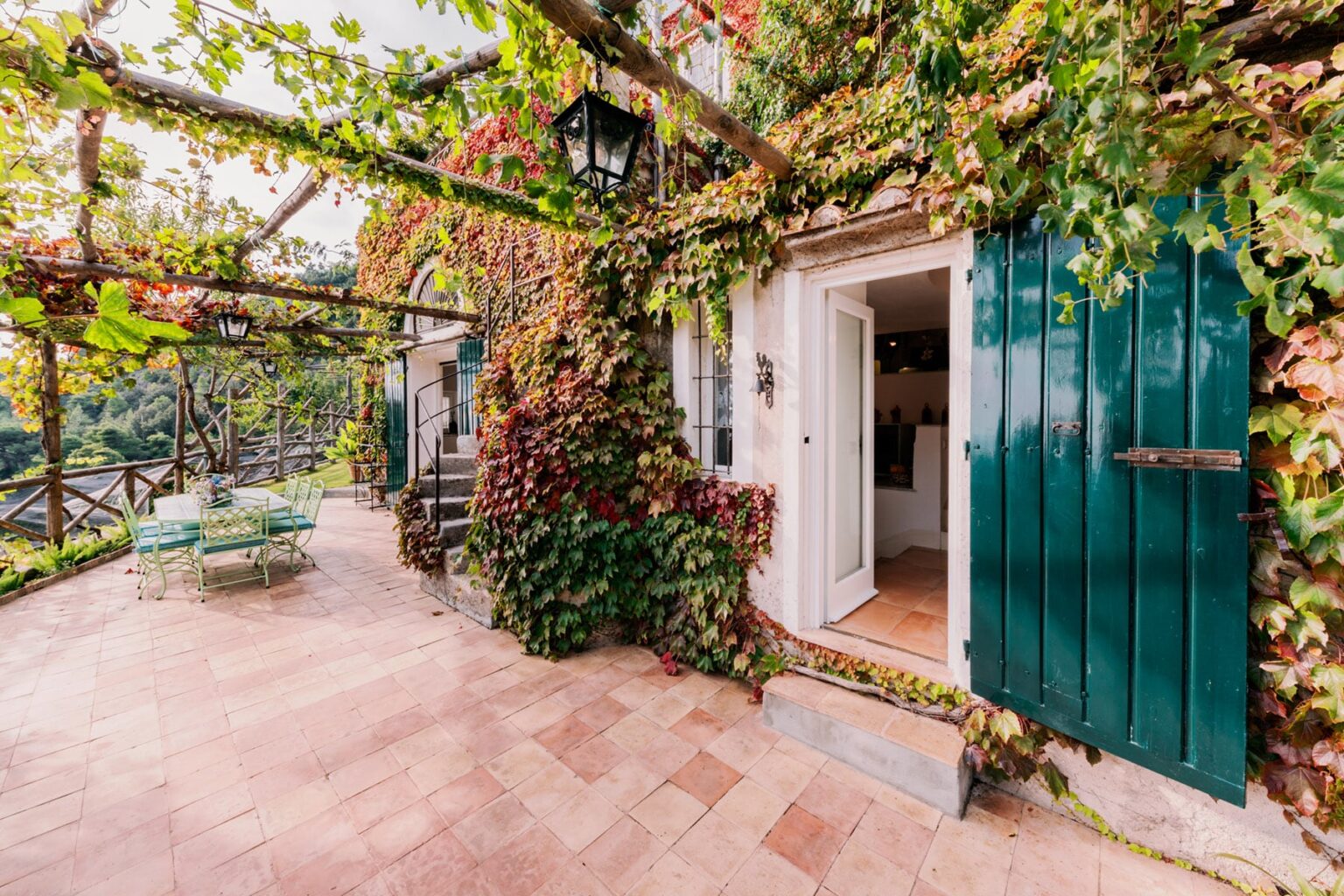 Charming exterior of a house with green wooden doors, surrounded by vibrant ivy and a patio area featuring outdoor seating. | Only on SopranoVillas