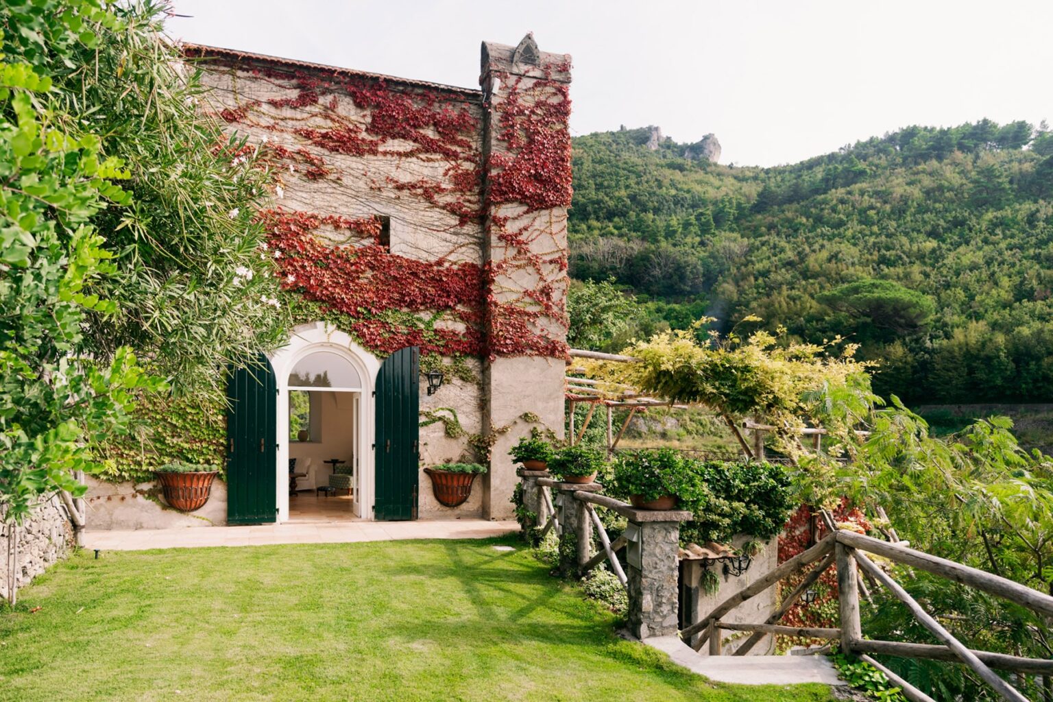 Charming exterior of a rustic villa covered in ivy, featuring arched doorways and large windows, surrounded by lush greenery and scenic hills in the background. | Only on SopranoVillas