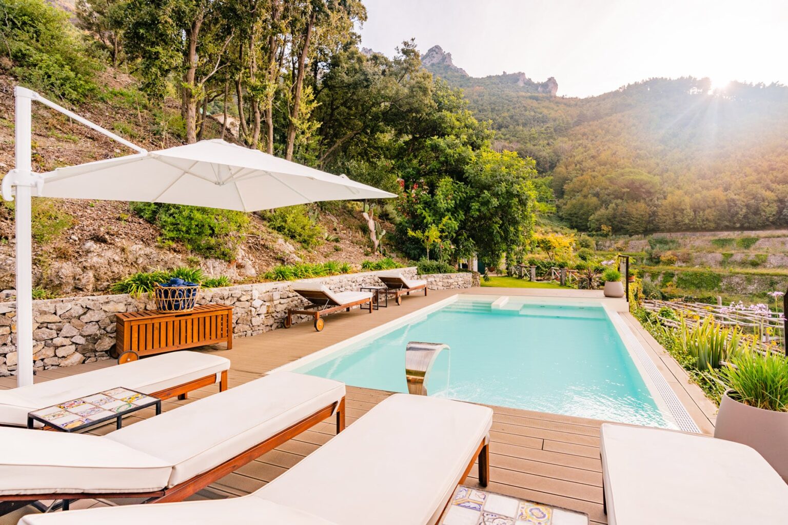 Luxurious outdoor pool area with sun loungers and a shaded umbrella, surrounded by lush greenery and hills, perfect for relaxation and leisure. | Only on SopranoVillas