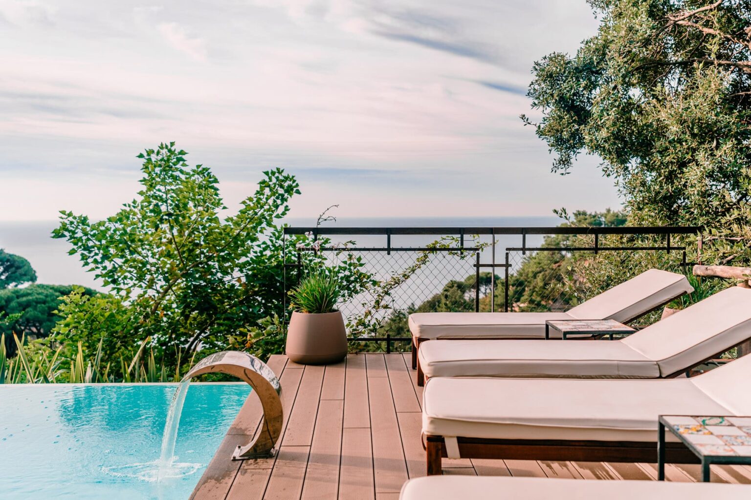 Luxurious outdoor pool area with comfortable lounge chairs, surrounded by greenery and offering a stunning view of the ocean under a serene sky. | Only on SopranoVillas