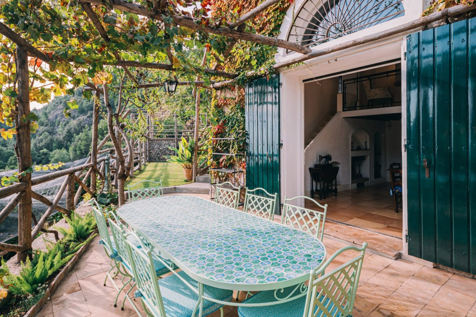 Outdoor dining area featuring a glass-topped table and green metal chairs, surrounded by lush greenery and vines, with a view of a house entrance and scenic landscape. Ideal for alfresco dining and gatherings. | Only on SopranoVillas