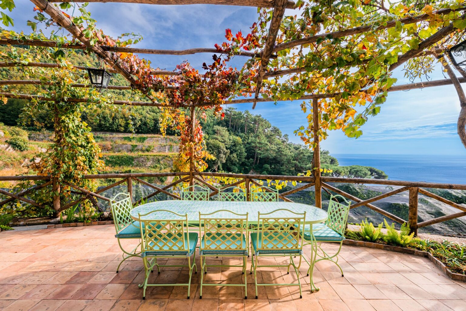 Outdoor dining area with a turquoise table and chairs, surrounded by lush greenery and vines, overlooking a scenic ocean view. | Only on SopranoVillas