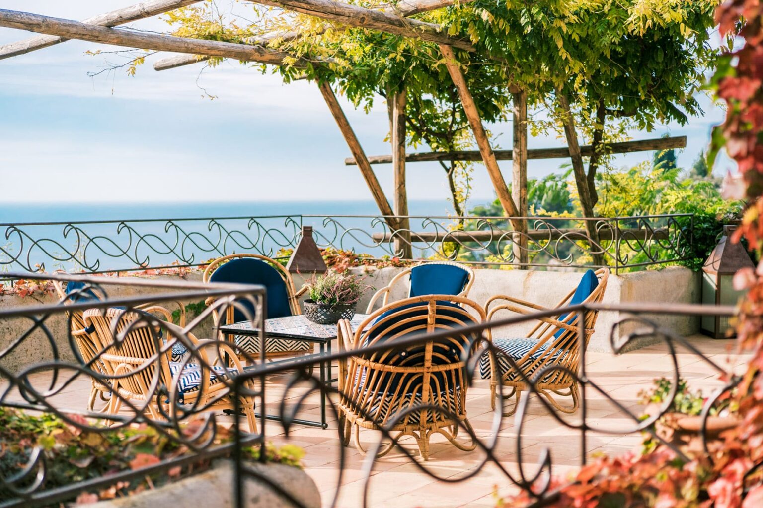 Cozy outdoor seating area with rattan chairs and a table overlooking the ocean, surrounded by greenery and a decorative railing. Perfect for relaxation and enjoying scenic views. | Only on SopranoVillas