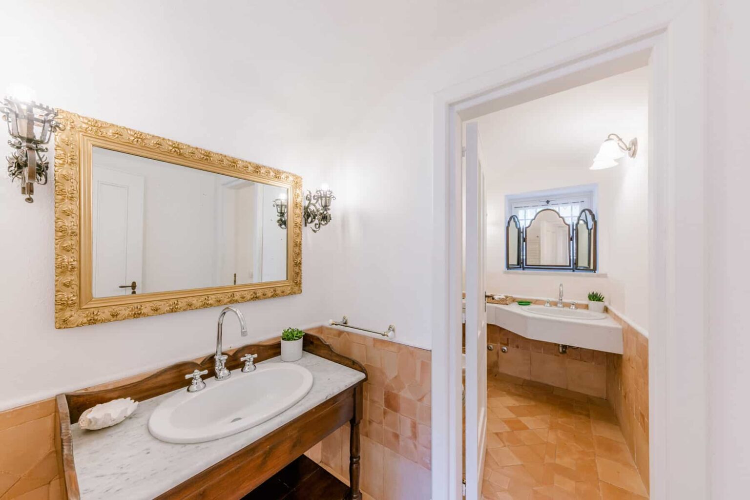 Elegant bathroom interior featuring a marble countertop with a dual sink, ornate golden mirror, and stylish lighting fixtures. The design includes a modern aesthetic with warm tones and natural light. | Only on SopranoVillas