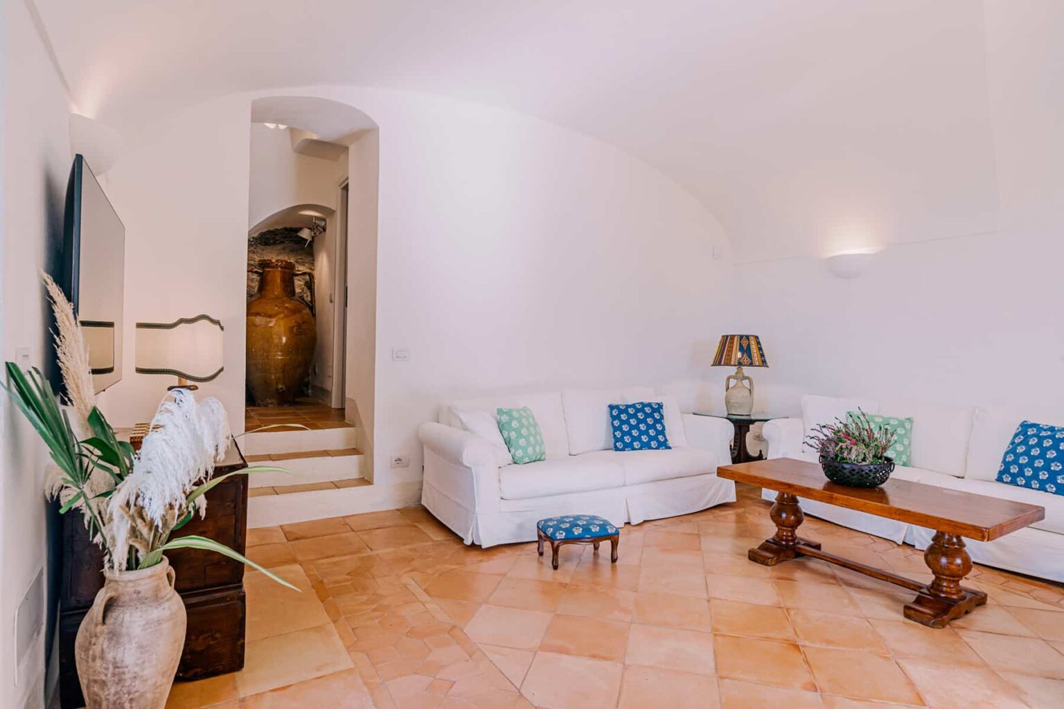 Bright and airy living room featuring a white sectional sofa with blue accent pillows, a wooden coffee table, and decorative plants. The space includes a staircase leading to an archway, enhancing the open and inviting atmosphere. Ideal for showcasing modern interior design. | Only on SopranoVillas