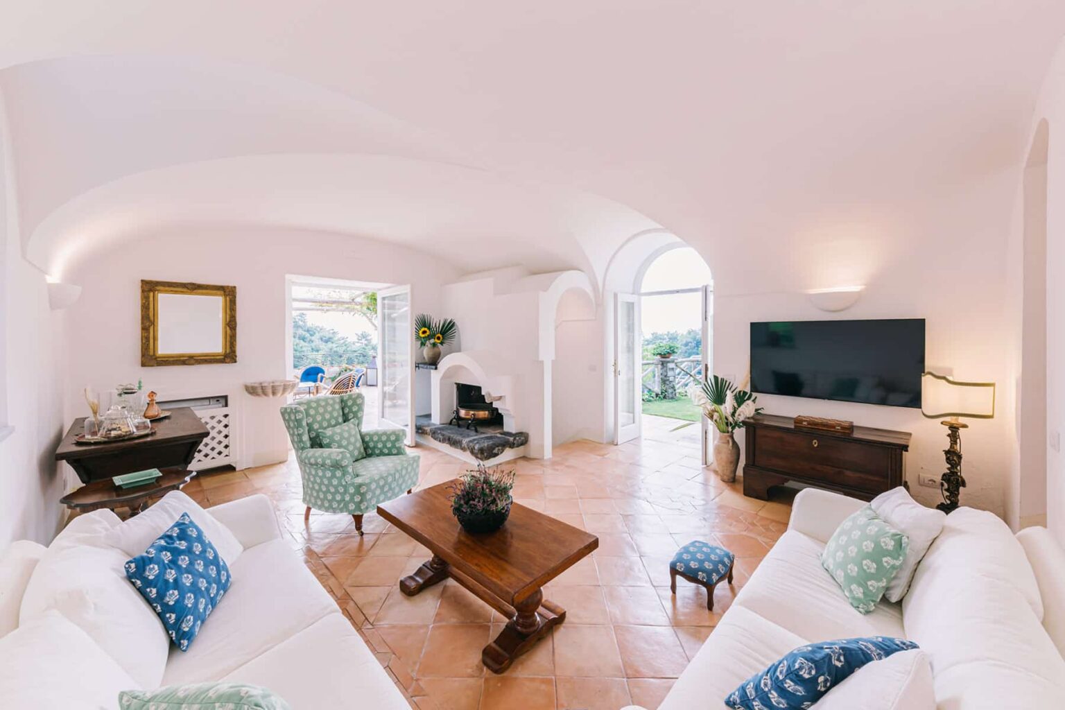 Bright and inviting modern living room featuring white sofas, a wooden coffee table, and stylish armchairs. The room includes a fireplace, a large flat-screen TV, and large windows that open to a garden view, creating a cozy and serene atmosphere. | Only on SopranoVillas