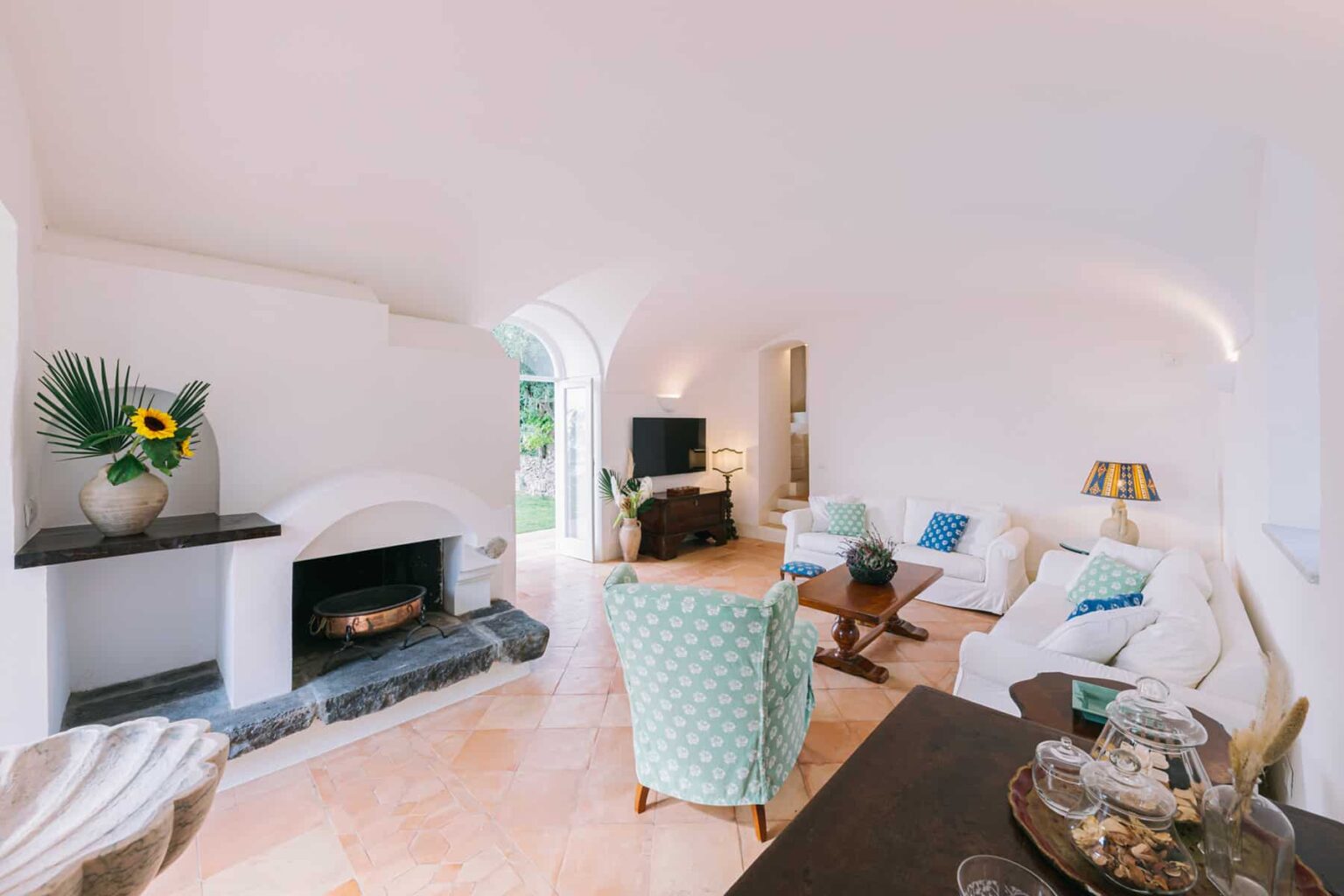 Cozy living room featuring a modern fireplace, light-colored furniture, and large windows allowing natural light, accented with decorative plants and a stylish lamp. | Only on SopranoVillas